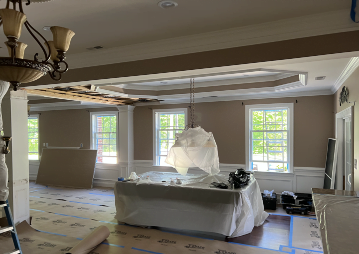 A living room is prepped for painting with floor coverings taped down and furniture covered in the middle of the room