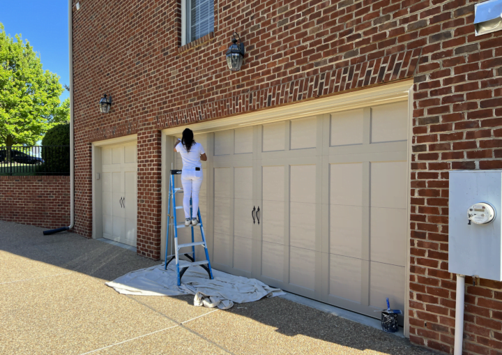 A woman on a ladder wearing a white painting outfit paints the trim of a garage