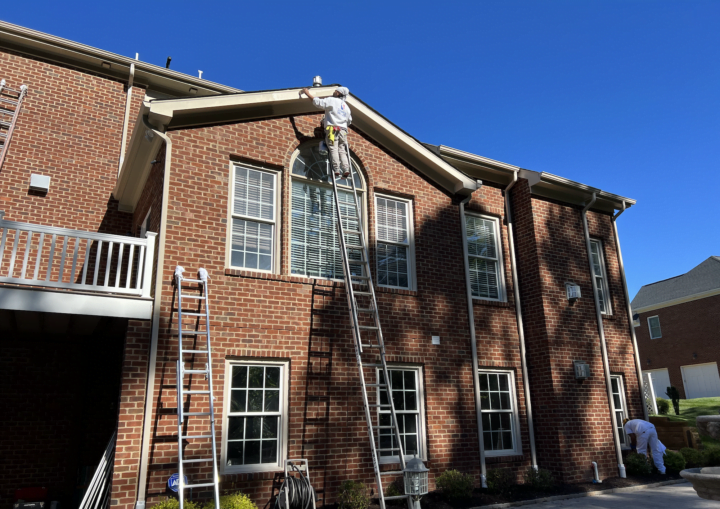 Men on tall ladders work to paint the trim on the outside of a brick house