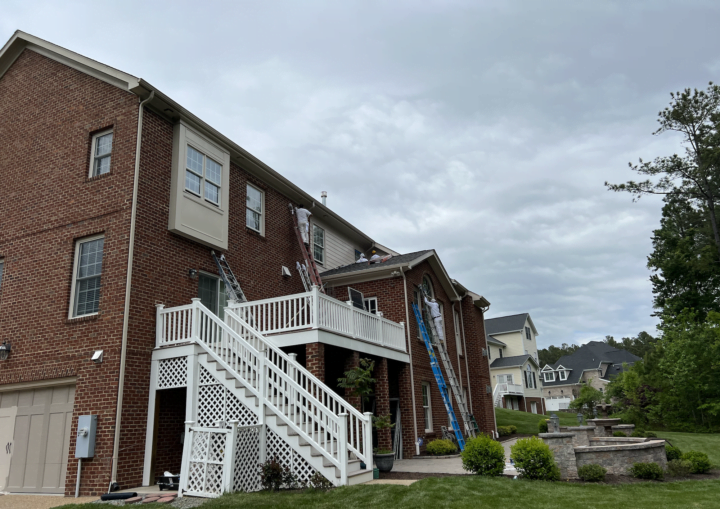 Men on tall ladders work to paint the trim on the outside of a brick house