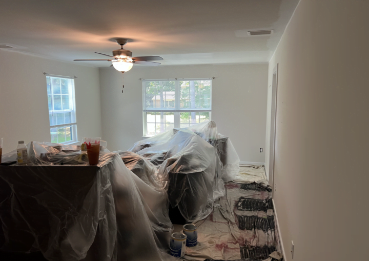 A room is prepped for painting with the furniture moved to the center of the room and covered with plastic