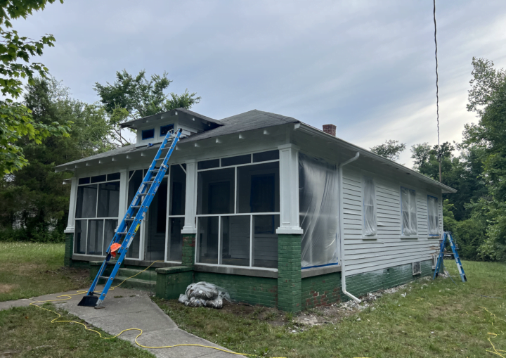 A blue ladder leans against a white bungalow as it is in the process of being painted